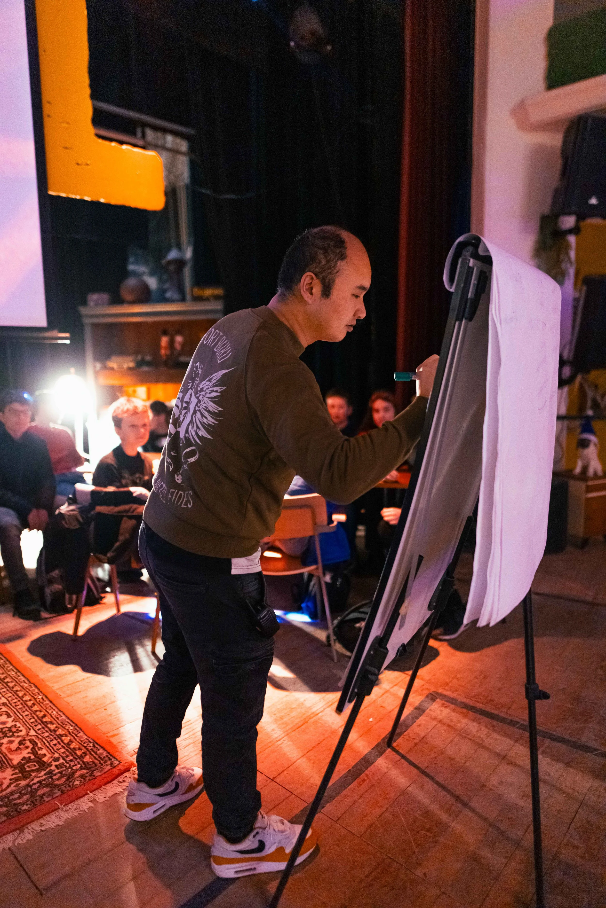 A man writing on a flip chart in front of an audience in a dimly lit room with a projector screen and seated people.