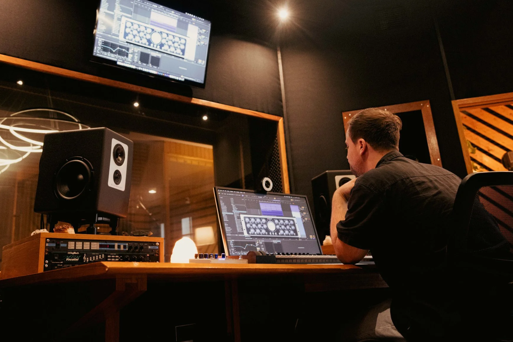Iemand aan het werk in een muziekstudio met een DAW op laptop en monitor, studiospeakers op het bureau en een akoestisch behandelde ruimte.