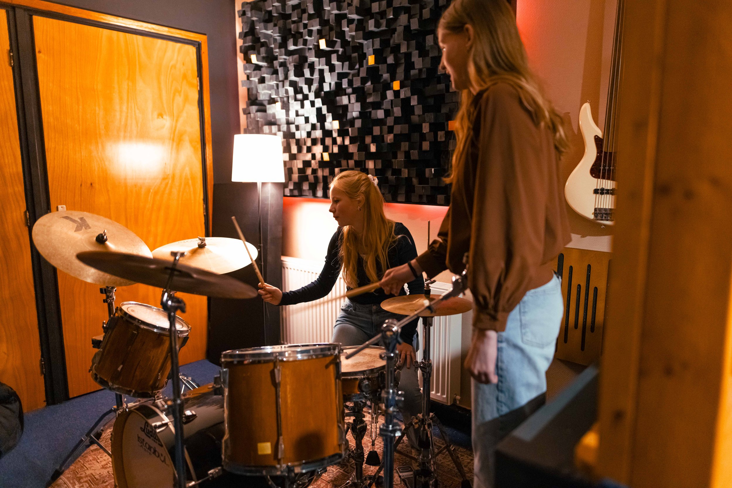 Twee vrouwen in een muziekstudio, de één achter een drumstel met een drumstok, de ander ernaast, omringd door instrumenten en akoestische panelen.